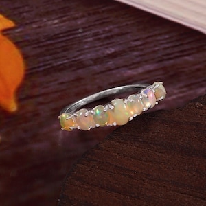 Puede incluir: Un anillo de plata con una fila de ópalos ovalados. Los ópalos muestran colores como blanco, amarillo, verde y rosa. El anillo está sobre una superficie de madera oscura. Es una pieza de joyería.