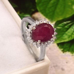 Puede incluir: Anillo de plata con un gran rubí ovalado, rodeado de piedras claras más pequeñas. El rubí es de un rojo intenso y el anillo tiene un acabado plateado pulido. Diseño elegante.