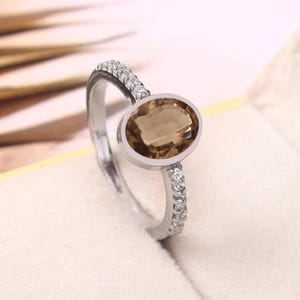 May include: A silver ring with a large, oval-cut, light brown gemstone. The band is embellished with small, clear gemstones. The ring is set against a soft, off-white background, accentuating the jewellery's design.