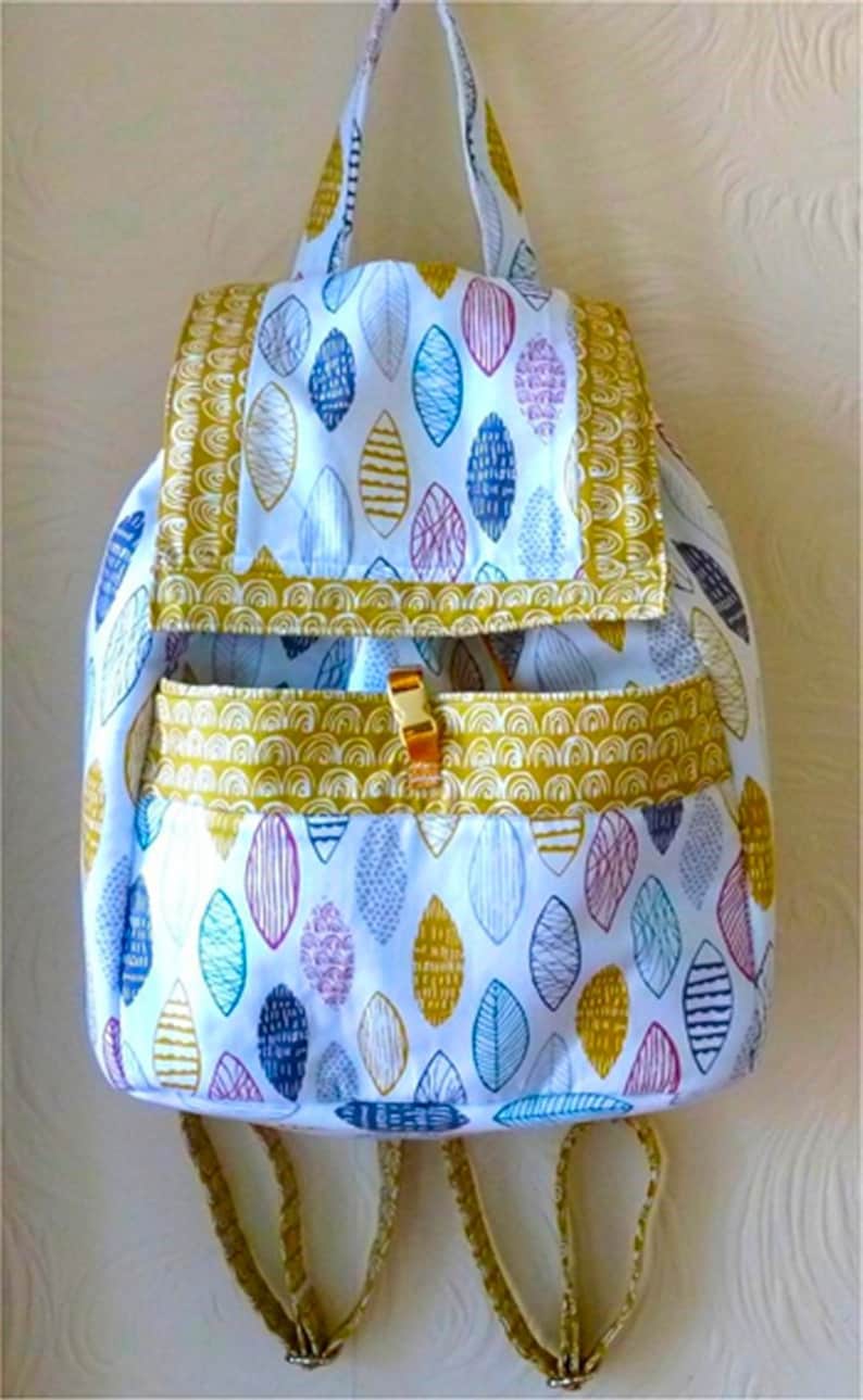 Pu&ograve; includere: Uno zaino bianco con un motivo a foglie gialle, blu e rosa. Lo zaino ha una patta con un motivo giallo, bianco e blu. Lo zaino ha due spalline regolabili.