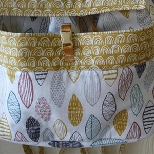 Pu&ograve; includere: Uno zaino in tessuto bianco con una tasca a motivo giallo e bianco. La tasca ha una chiusura in metallo dorato. Il tessuto ha un motivo di foglie colorate.
