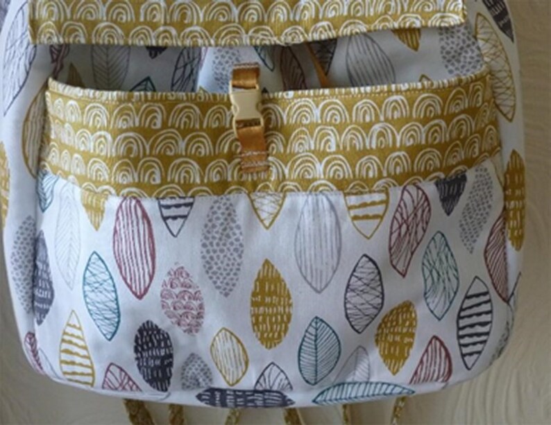 Pu&ograve; includere: Uno zaino in tessuto bianco con una tasca a motivo giallo e bianco. La tasca ha una chiusura in metallo dorato. Il tessuto ha un motivo di foglie colorate.