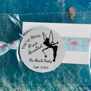 May include: A clear plastic bag contains a light blue bow, a round silver sticker with a Tinkerbell silhouette and text, and a light blue elastic band with pink flamingoes. Two seahorse pins are also visible.