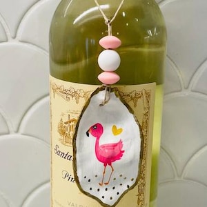 May include: A clear glass wine bottle with a gold foil neck, featuring a decorative oyster shell ornament tied with twine. The shell is painted with a pink flamingo, a yellow heart, and black dots, accented by two pink and one white silicone beads.