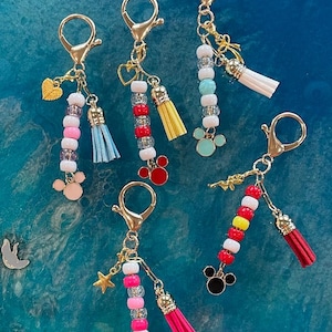May include: A collection of gold-toned keychains with colorful beaded accents and small tassels. Each keychain features a different color scheme, including pink, blue, red, and yellow beads, and is adorned with charms like hearts, stars, and Mickey Mouse silhouettes.