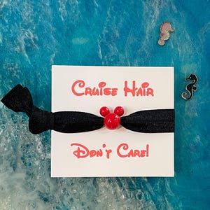 May include: Black hair tie with a red Mickey Mouse head charm. The hair tie is on a white card with the text "Cruise Hair Don't Care!"