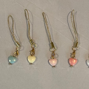May include: Six heart-shaped charms in pastel colors, each with a gold-colored clasp and cord. The hearts are in shades of purple, blue, yellow, pink, and white. The charms are likely for a phone, keychain, or other accessory.