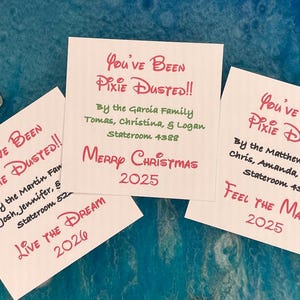 May include: White cards with red and green text that reads "You've Been Pixie Dusted!!" and "Merry Christmas 2025". The cards also include names and dates. The background is a blue, textured surface.