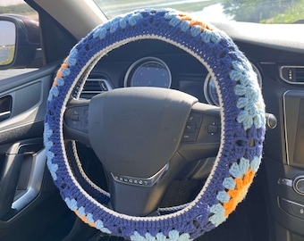 May include: A blue, white, and orange crocheted steering wheel cover with a flower pattern. The cover is on a black steering wheel with the word "PEUGEOT" in white.