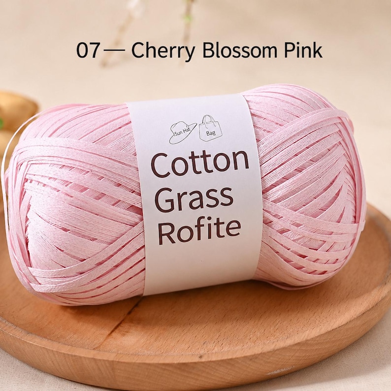 May include: A ball of light pink ribbon yarn labelled 07 Cherry Blossom Pink. The white paper band features the text Cotton Grass Rofite and icons of a sun hat and a bag. The yarn has a flat, tape-like texture and is wound into a compact oval shape.