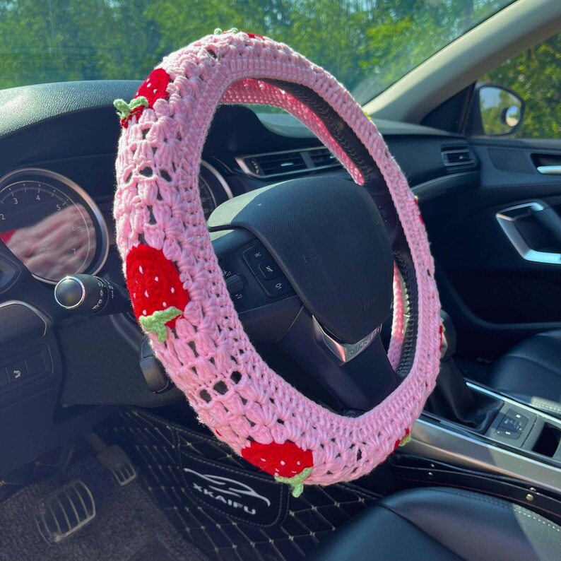 May include: A pink crocheted steering wheel cover adorned with red strawberries and green leaves. The cover is installed on a black steering wheel inside a car. The strawberry design adds a touch of whimsy to the vehicle's interior.
