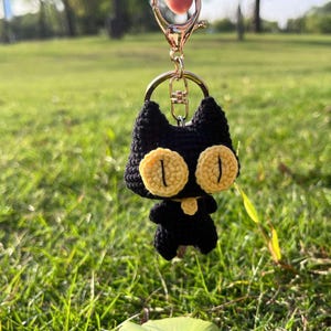 May include: Black crocheted cat keychain with yellow eyes. The keychain is attached to a silver key ring.