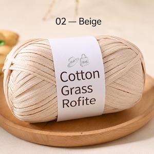 May include: A ball of beige ribbon yarn with a white paper label featuring the text 02 - Beige, Cotton Grass Rofite, and small line drawings of a hat and a bag. The yarn is wound into an oval shape and has a flat, smooth, and matte texture.