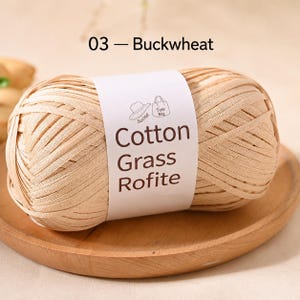May include: A ball of light beige raffia-style yarn labelled 03 Buckwheat. The white paper band features the text Cotton Grass Rofite, along with small line-art icons of a sun hat and a tote bag, indicating the intended use for these items.