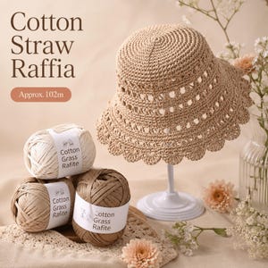 May include: A beige crocheted bucket hat with a scalloped edge sits on a white stand next to three balls of yarn labelled Cotton Grass Rafite. The text Cotton Straw Raffia and Approx. 102m is visible. The yarn is in shades of cream and light brown.