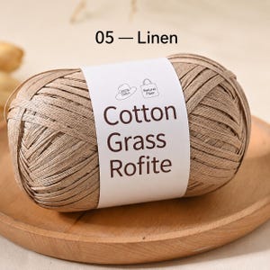 May include: A ball of light brown, flat, ribbon-like yarn labelled 05 - Linen. The white paper band features the text Cotton Grass Rofite, along with small icons of a hat and a tote bag, indicating 100% cotton and natural fibre content.