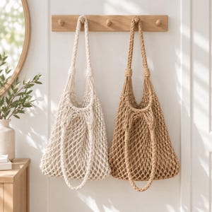 May include: Two macrame tote bags hanging from a wooden wall rack. One bag is off-white and the other is tan. Both bags feature an open net design, braided shoulder straps, and knotted detailing at the top where the straps meet the main body.