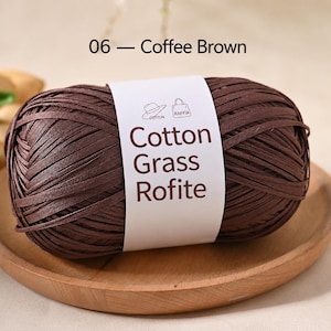 May include: A ball of coffee brown yarn wrapped in a white paper label with the text 06 - Coffee Brown and Cotton Grass Rofite. The label features small line art icons of a hat and a handbag, indicating the material is suitable for crafting.