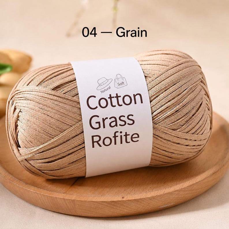 May include: A ball of light brown, flat ribbon-like yarn labelled 04 - Grain. The white paper band features the text Cotton Grass Rofite, along with small icons of a hat and a bag, and the words Natural and Soft printed in dark brown ink.