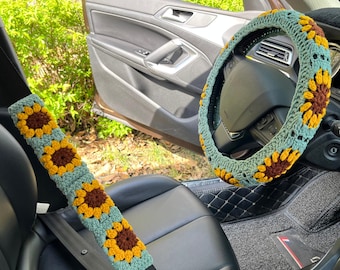 May include: A car interior with a crocheted sunflower steering wheel cover and a crocheted sunflower seatbelt strap. The steering wheel cover is teal and brown with yellow sunflowers. The seatbelt strap is teal and yellow with brown sunflowers.