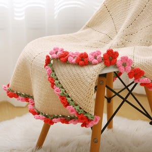 May include: A cream-colored crocheted blanket draped over a wooden chair. The blanket is adorned with a border of red, pink, and green crocheted flowers. The chair has a light wood finish and black metal supports.