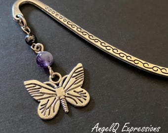 Può includere: Un segnalibro in metallo argentato a forma di gancio con un motivo a volute inciso lungo l'asta. Un ciondolo pendente include una piccola perlina nera, una perlina rotonda viola e una farfalla argentata. Il testo AngelQ Expressions è visibile.