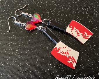 Può includere: Un paio di orecchini con un design a coltello da macellaio rosso e bianco con schizzi di sangue rosso. Gli orecchini sono attaccati a ganci d'argento con un ciondolo a forma di cuore rosso e iridescente.