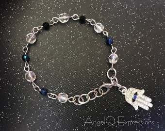 Può includere: Un braccialetto a catena d'argento con perline trasparenti e nere, e un ciondolo a forma di mano d'argento con un occhio turco blu. Il braccialetto ha il testo "AngelQExpressions" in basso.