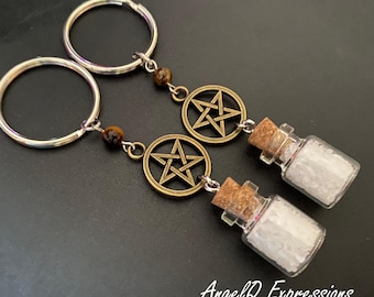 Può includere: Due portachiavi con un anello in metallo argentato, una perla occhio di tigre marrone, un ciondolo a pentagramma color bronzo e una piccola bottiglia di vetro trasparente riempita di cristalli di sale bianco, sigillata con un tappo di sughero.