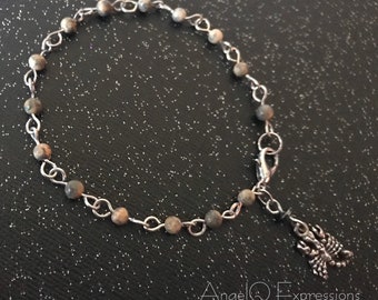 Può includere: Un braccialetto a catena d'argento con perline macchiate di marrone e grigio e un ciondolo a forma di scorpione d'argento.