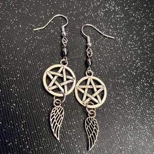 Może przedstawiać: Para srebrnych kolczyk&oacute;w wiszących. Każdy kolczyk ma zawieszkę w kształcie pentagramu, sznur ciemnych koralik&oacute;w i małą zawieszkę w kształcie skrzydła. Tło to ciemna, błyszcząca powierzchnia.