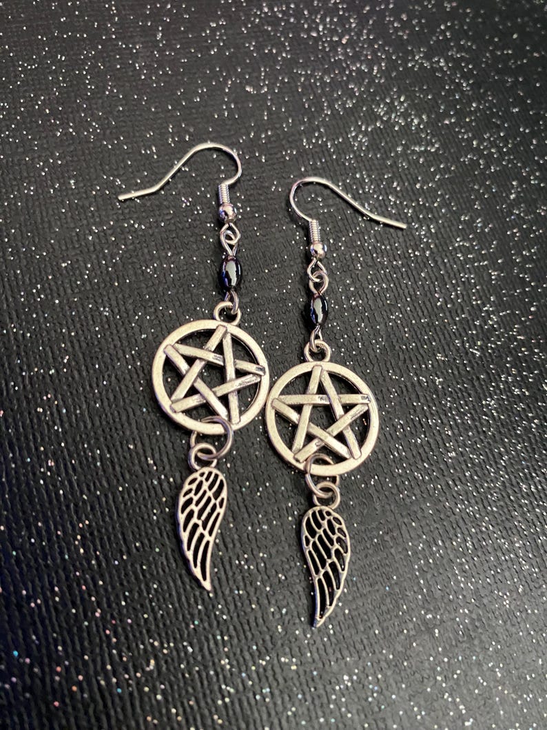Może przedstawiać: Para srebrnych kolczyk&oacute;w wiszących. Każdy kolczyk ma zawieszkę w kształcie pentagramu, sznur ciemnych koralik&oacute;w i małą zawieszkę w kształcie skrzydła. Tło to ciemna, błyszcząca powierzchnia.