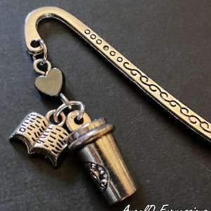 May include: A silver-toned metal hook bookmark featuring a decorative scroll pattern along the stem. Attached to the hook are three charms: a small solid heart, an open book with visible pages, and a coffee cup. The text AngelQ Expressions is visible.