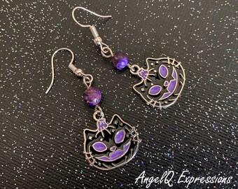 Può includere: Un paio di orecchini pendenti con un viso di gatto nero con accenti viola e una bocca cucita. Gli orecchini sono realizzati in metallo argentato e perline viola.