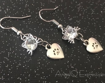 Può includere: Orecchini pendenti color argento. Ogni orecchino presenta un ciondolo a forma di gatto con un centro di cristallo trasparente, un ciondolo a forma di cuore con impronte di zampe e una chiusura a gancio. Sfondo nero con macchie bianche. Testo: Angel Q Expressions.