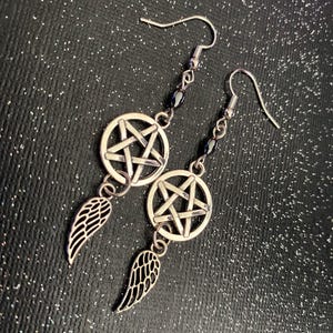 Może przedstawiać: Srebrne kolczyki z pentagramem w okręgu, czarnymi koralikami i zawieszką w kształcie skrzydła. Zapięcie na haczyk. Ciemne, błyszczące tło. Odpowiednie dla gotyckiej estetyki.