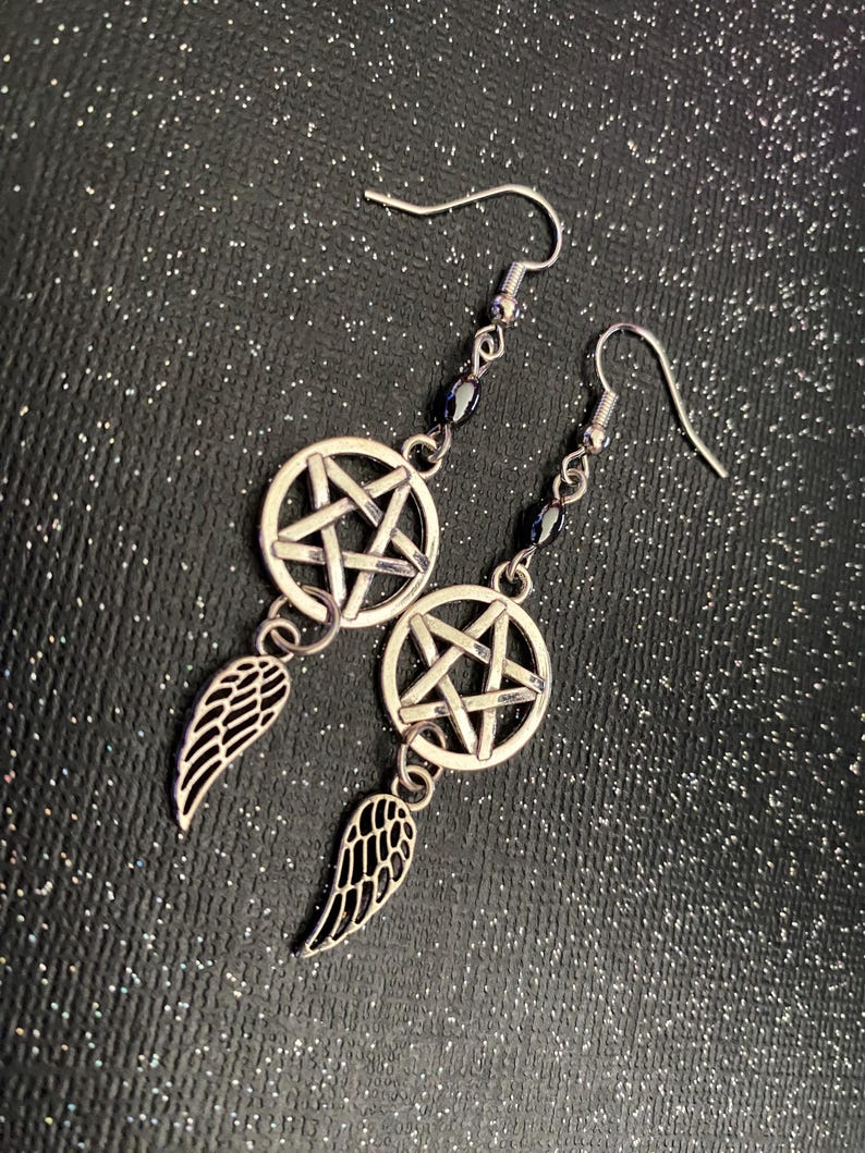 Może przedstawiać: Srebrne kolczyki z pentagramem w okręgu, czarnymi koralikami i zawieszką w kształcie skrzydła. Zapięcie na haczyk. Ciemne, błyszczące tło. Odpowiednie dla gotyckiej estetyki.