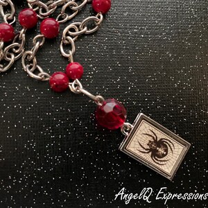 May include: A silver chain necklace with red beads and a silver charm featuring a black spider on a white web. The charm is rectangular and has a silver frame. The necklace is on a black background with glitter.