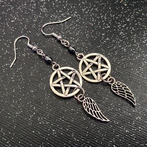 Może przedstawiać: Srebrne kolczyki z wisiorkiem w kształcie pentagramu i zawieszką w kształcie skrzydła. Kolczyki mają ciemny akcent z koralik&oacute;w i zapięcie na haczyk. Prezentowane są na czarnej, błyszczącej powierzchni.