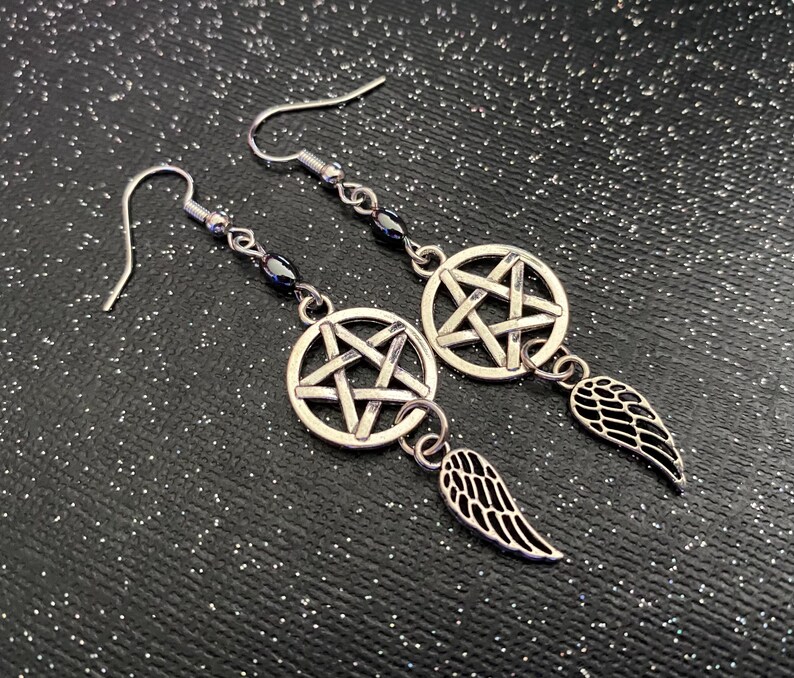Może przedstawiać: Srebrne kolczyki z wisiorkiem w kształcie pentagramu i zawieszką w kształcie skrzydła. Kolczyki mają ciemny akcent z koralik&oacute;w i zapięcie na haczyk. Prezentowane są na czarnej, błyszczącej powierzchni.