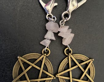 Può includere: Due chiusure a moschettone color argento attaccate a perline di pietra rosa pallido, irregolari e levigate, collegate a ciondoli a forma di pentagramma in metallo color bronzo. Il testo "AngelQ Expressions" è stampato in bianco nell'angolo in basso a destra.