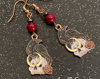 Può includere: Orecchini pendenti color oro con un design di corvo nero appollaiato su un teschio, una luna crescente e una rosa. Gli orecchini hanno accenti di perline rosse e nere. Il testo "AngelQ Expressions" è in basso a destra.