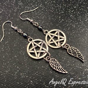 Może przedstawiać: Srebrne kolczyki wiszące. Każdy kolczyk posiada pentagram w okręgu, z małym urokiem skrzydła. Kolczyki mają zapięcie na haczyk i są połączone łańcuszkiem z ciemnymi koralikami. Tło jest czarne z brokatem.