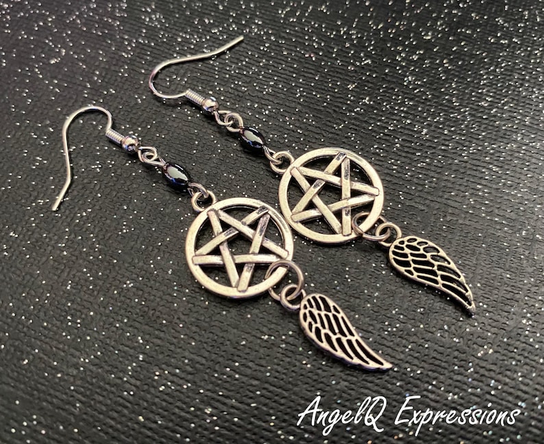 Może przedstawiać: Srebrne kolczyki wiszące. Każdy kolczyk posiada pentagram w okręgu, z małym urokiem skrzydła. Kolczyki mają zapięcie na haczyk i są połączone łańcuszkiem z ciemnymi koralikami. Tło jest czarne z brokatem.