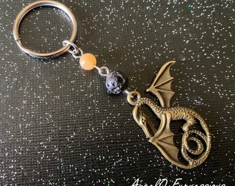 Può includere: Un portachiavi a forma di drago in bronzo con un anello portachiavi in argento e perline. Le perline sono arancioni e nere. Il drago è arrotolato con le ali spiegate. Il portachiavi è su uno sfondo nero scintillante.