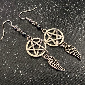 Może przedstawiać: Srebrne kolczyki z pentagramem w okręgu i małym skrzydłem. Kolczyki mają zapięcie na haczyk i są umieszczone na ciemnym, błyszczącym tle.