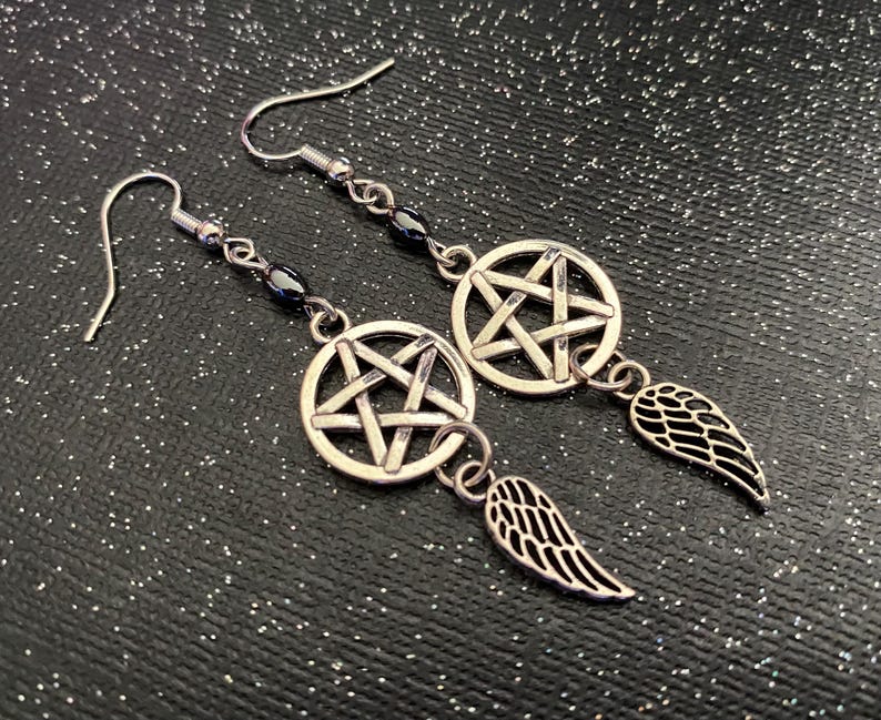 Może przedstawiać: Srebrne kolczyki z pentagramem w okręgu i małym skrzydłem. Kolczyki mają zapięcie na haczyk i są umieszczone na ciemnym, błyszczącym tle.