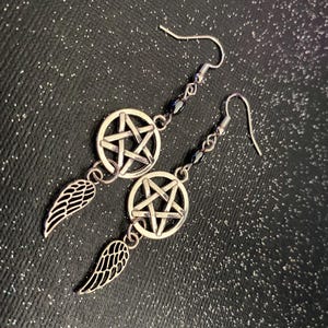 Może przedstawiać: Srebrne kolczyki wiszące z motywem pentagramu w okręgu i małym urokiem skrzydła. Kolczyki mają zapięcie na haczyk i są ozdobione małymi czarnymi koralikami. Tło to ciemna, błyszcząca powierzchnia.