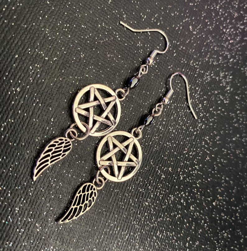 Może przedstawiać: Srebrne kolczyki wiszące z motywem pentagramu w okręgu i małym urokiem skrzydła. Kolczyki mają zapięcie na haczyk i są ozdobione małymi czarnymi koralikami. Tło to ciemna, błyszcząca powierzchnia.