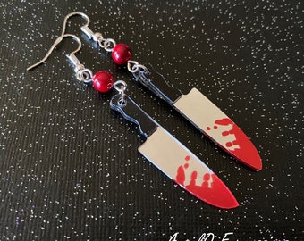 Può includere: Un paio di orecchini d'argento con perline rosse e un ciondolo a forma di coltello insanguinato. Il coltello è nero e argento con schizzi di vernice rossa.